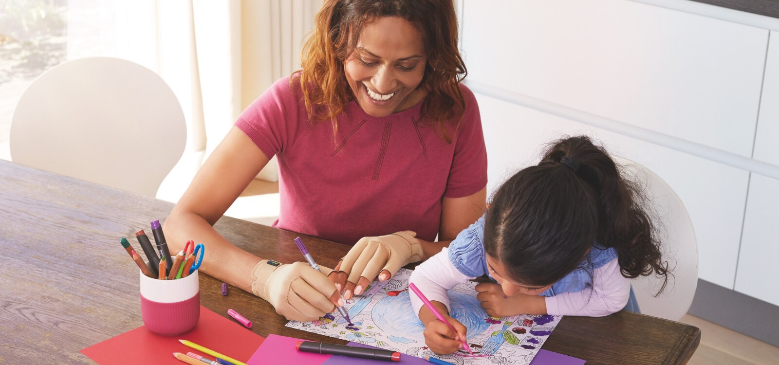 Mujer y niña coloreando un dibujo. La mujer lleva puestos los guantes para artritis Actimove Arthritis Care porque tiene molestias en el pulgar.