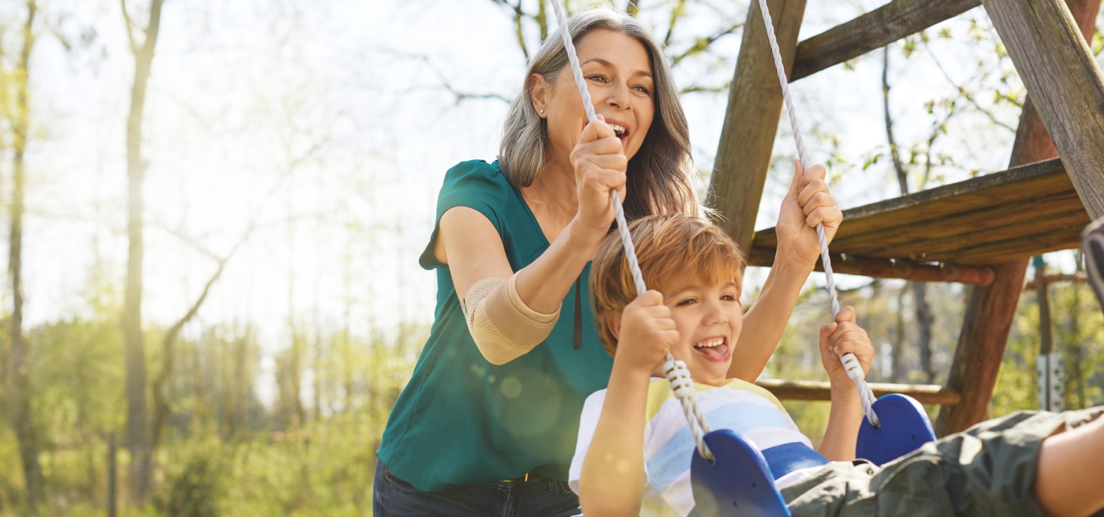 Mujer en el parque empujando a un niño en un columpio mientras usa la codera Actimove Everyday Supports para la epicondilitis. 