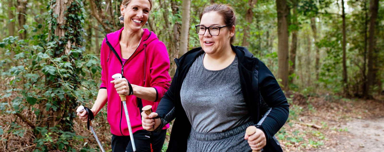 Two smiling women walking with poles in a wooded area.