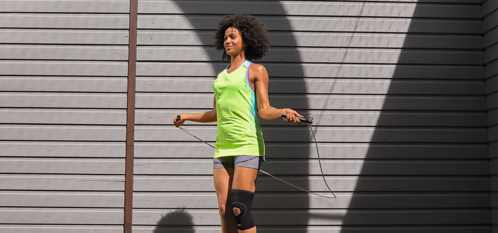 Ženska v športnih oblačilih s kolensko oporo na nogi med skakanjem čez kolebnico na prostem.